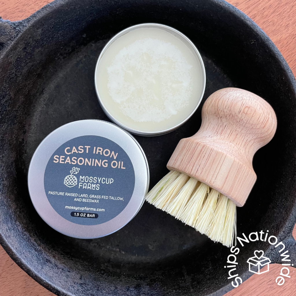 A cast iron pan sits on a wooden table. In the middle is an open tin jar of Mossycup Farms' Cast Iron Seasoning Oil and a bamboo bursh. In the corner a logo in white says Ships Nationwide, and a box with a heart coming out of the top sits in the middle.